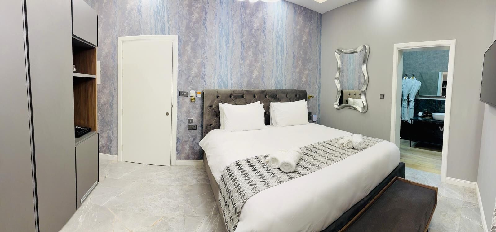 Una habitación de hotel moderna con una cama grande, un armario, un espejo y un baño visible a través de una puerta abierta.