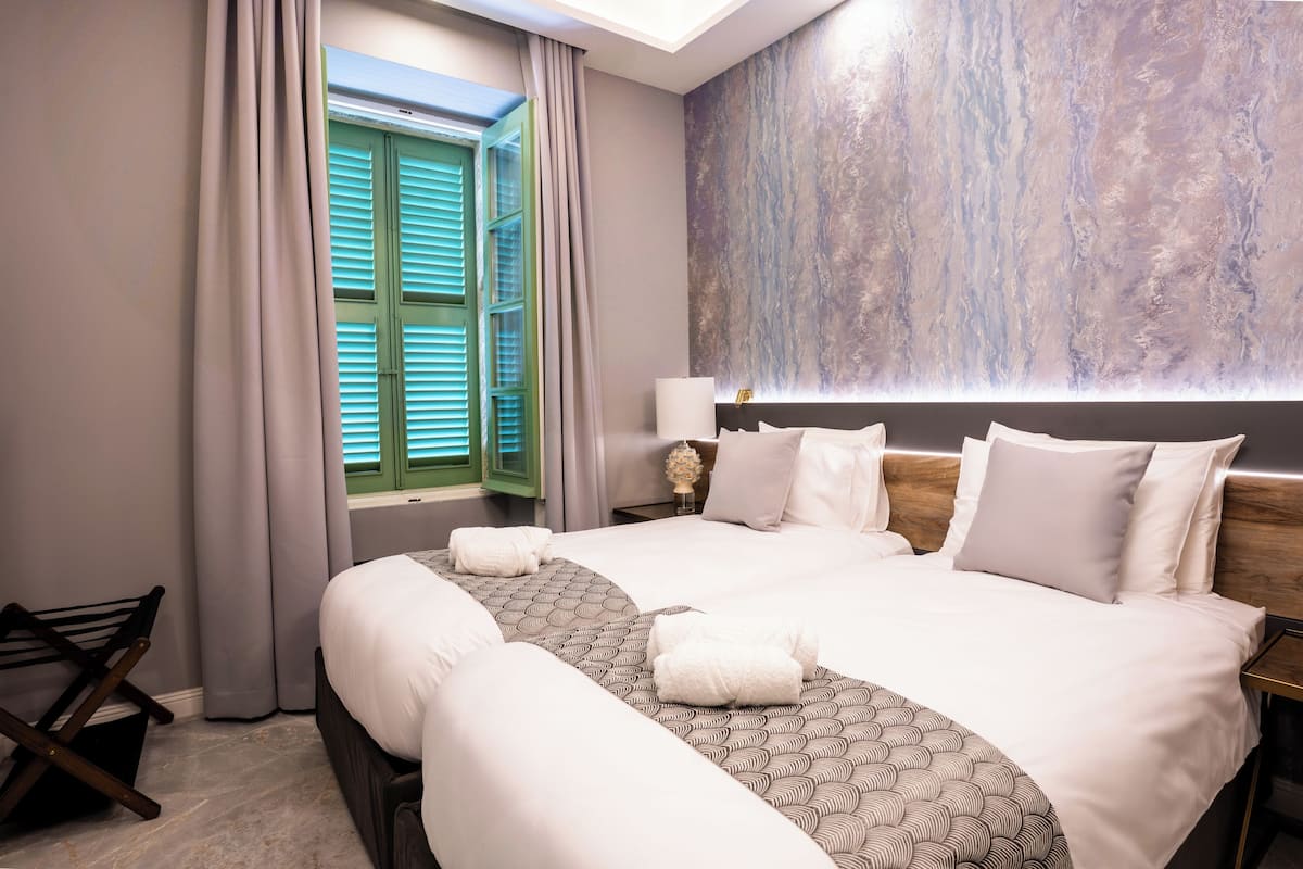 A hotel room with a large bed, a nightstand, a lamp, and a window with green shutters.