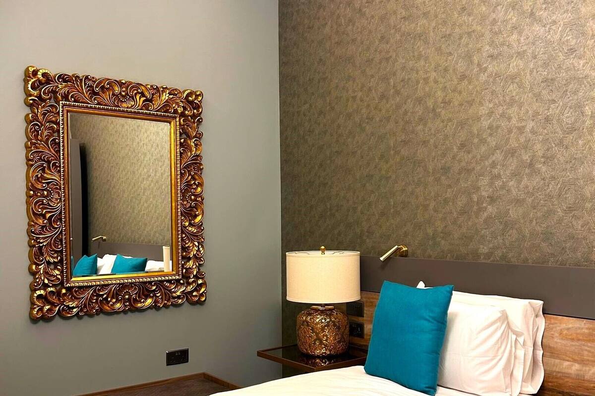 A hotel room with a large ornate mirror, a bed with blue and white bedding, a nightstand with a lamp, and a textured wall.