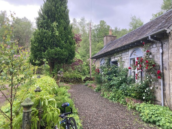Uma casa de pedra com um caminho de cascalho que a leva, cercada por vegetação exuberante e plantas floridas.