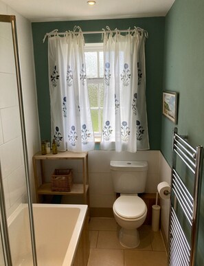 Uma casa de banho com sanita, banheira, janela com cortinas florais, prateleira com produtos de higiene pessoal e um quadro na parede.