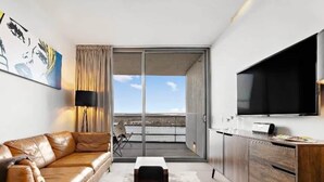 Une chambre d’hôtel moderne avec un canapé en cuir, un téléviseur à écran plat, un meuble en bois et une vue sur la ville, visible à travers une porte coulissante.