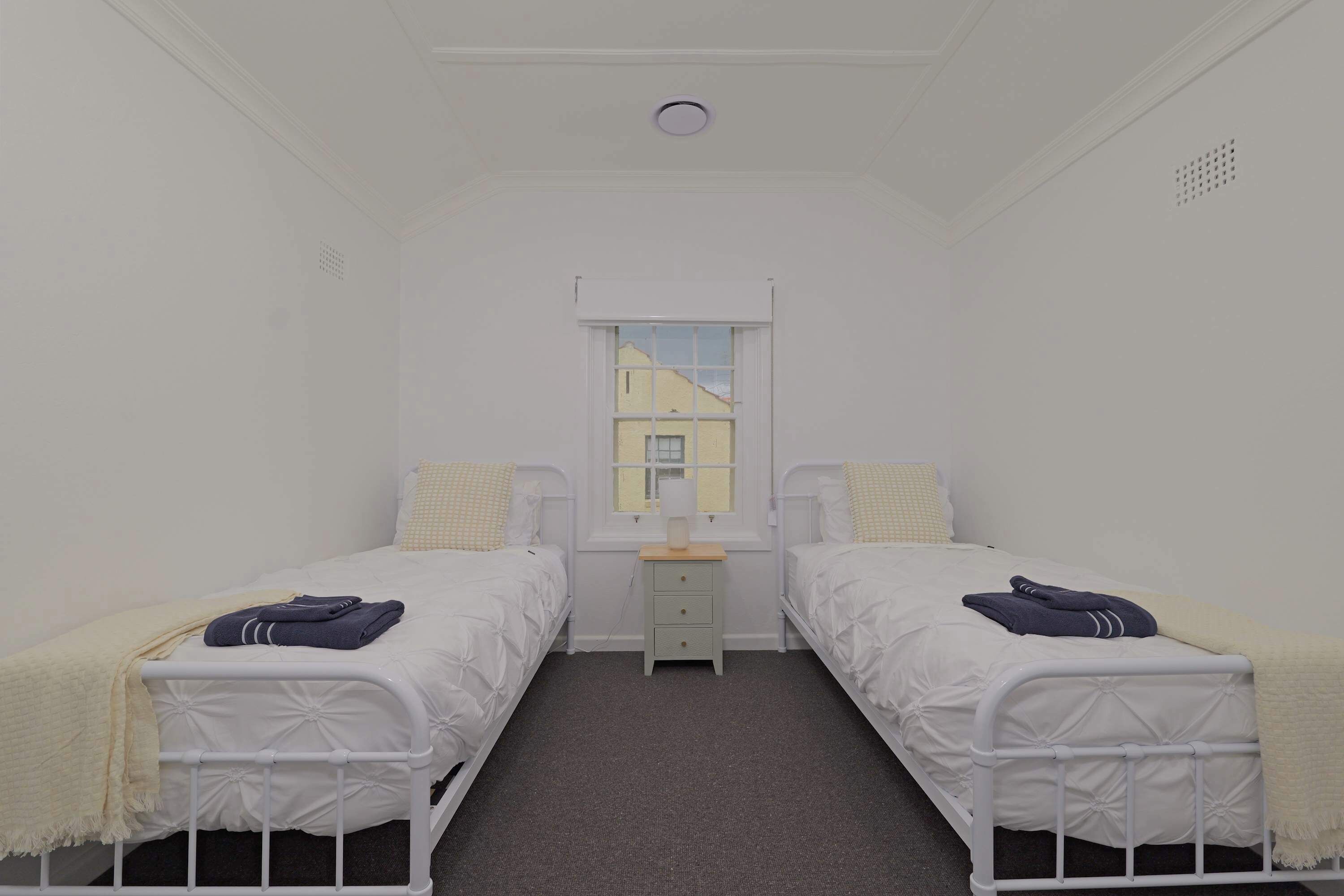 A small, clean room with two single beds, a small bedside table, a window, and a door.