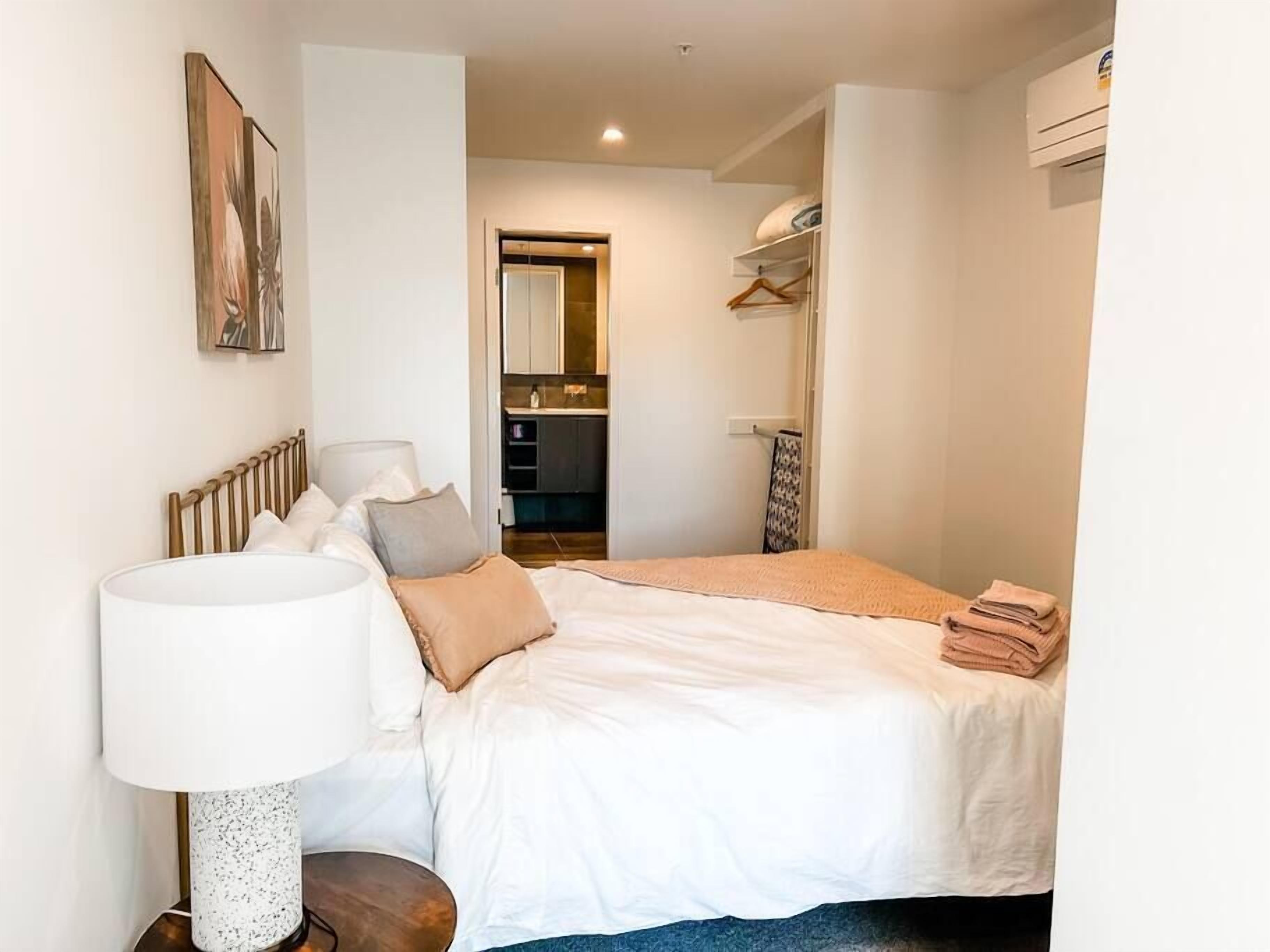 Quarto com cama, mesa de cabeceira, luminária e vista para a cozinha.