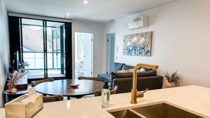 A modern kitchen with a large window, a dining area, and a living room with a sofa and a painting on the wall. - Luxurious Apartment in Canberra CBD (Reid)