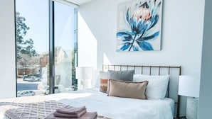 A modern bedroom with a large bed, a view of the street, and a painting on the wall. - Luxurious Apartment in Canberra CBD (Reid)