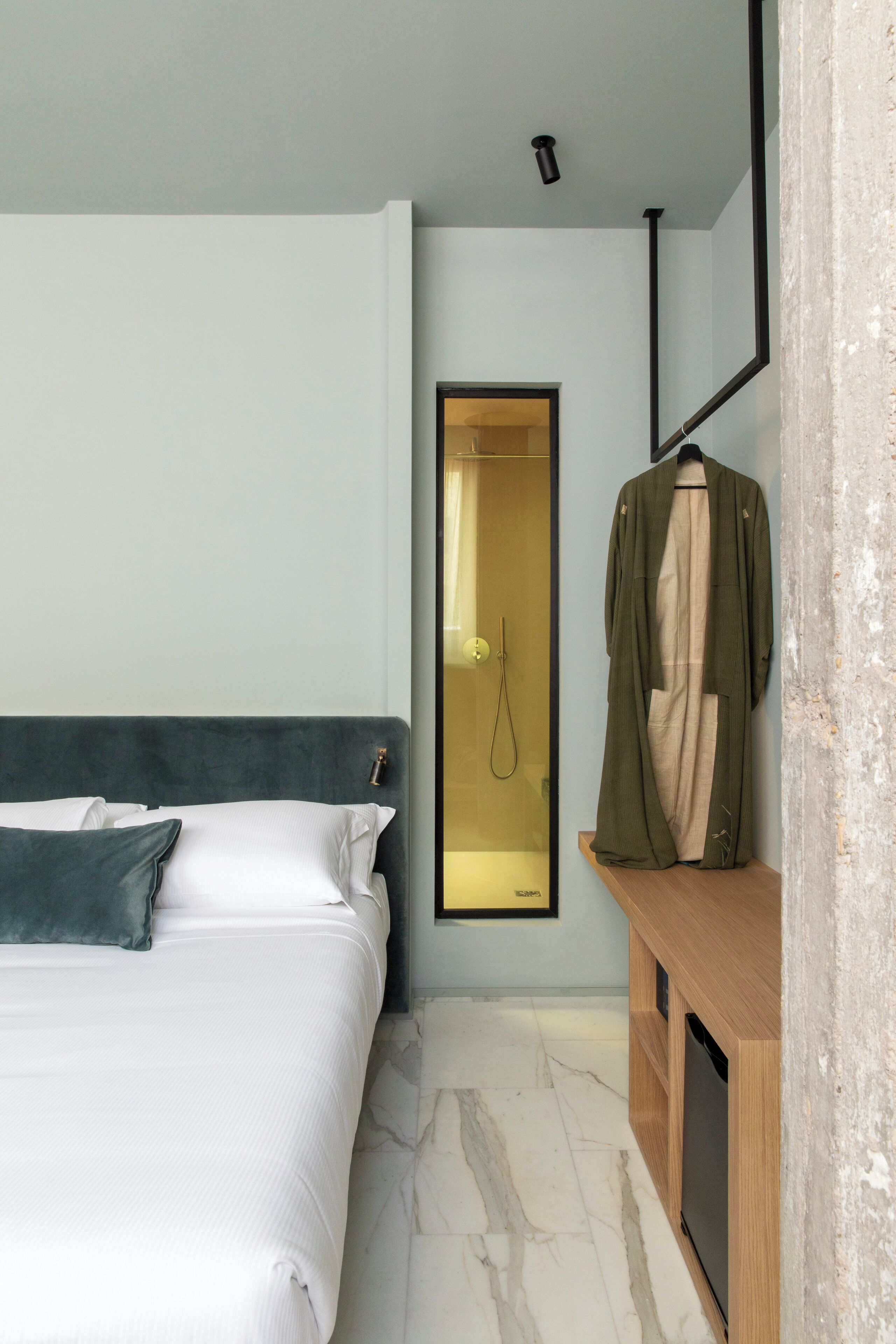 Quarto de hotel com cama, mesa de cabeceira de madeira, espelho e chuveiro.