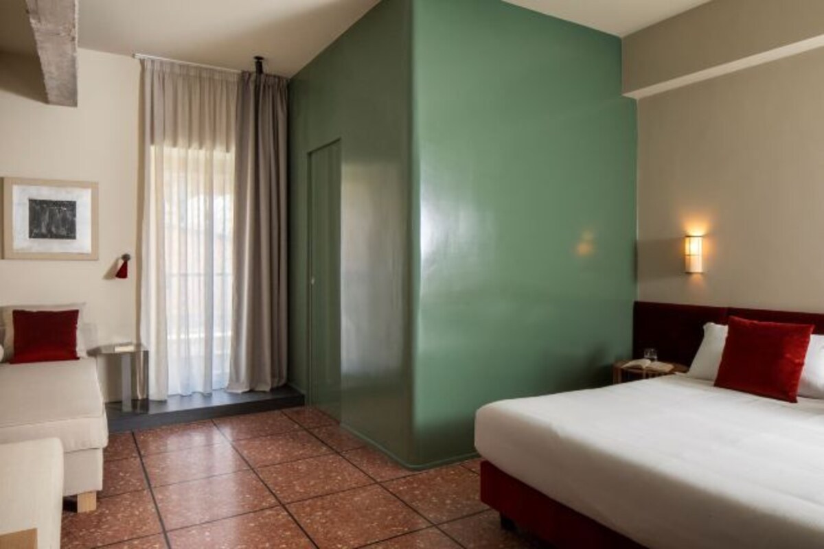 Une chambre d’hôtel avec un lit, une petite table, une chaise, une fenêtre avec des rideaux et un mur muni d’un interrupteur.