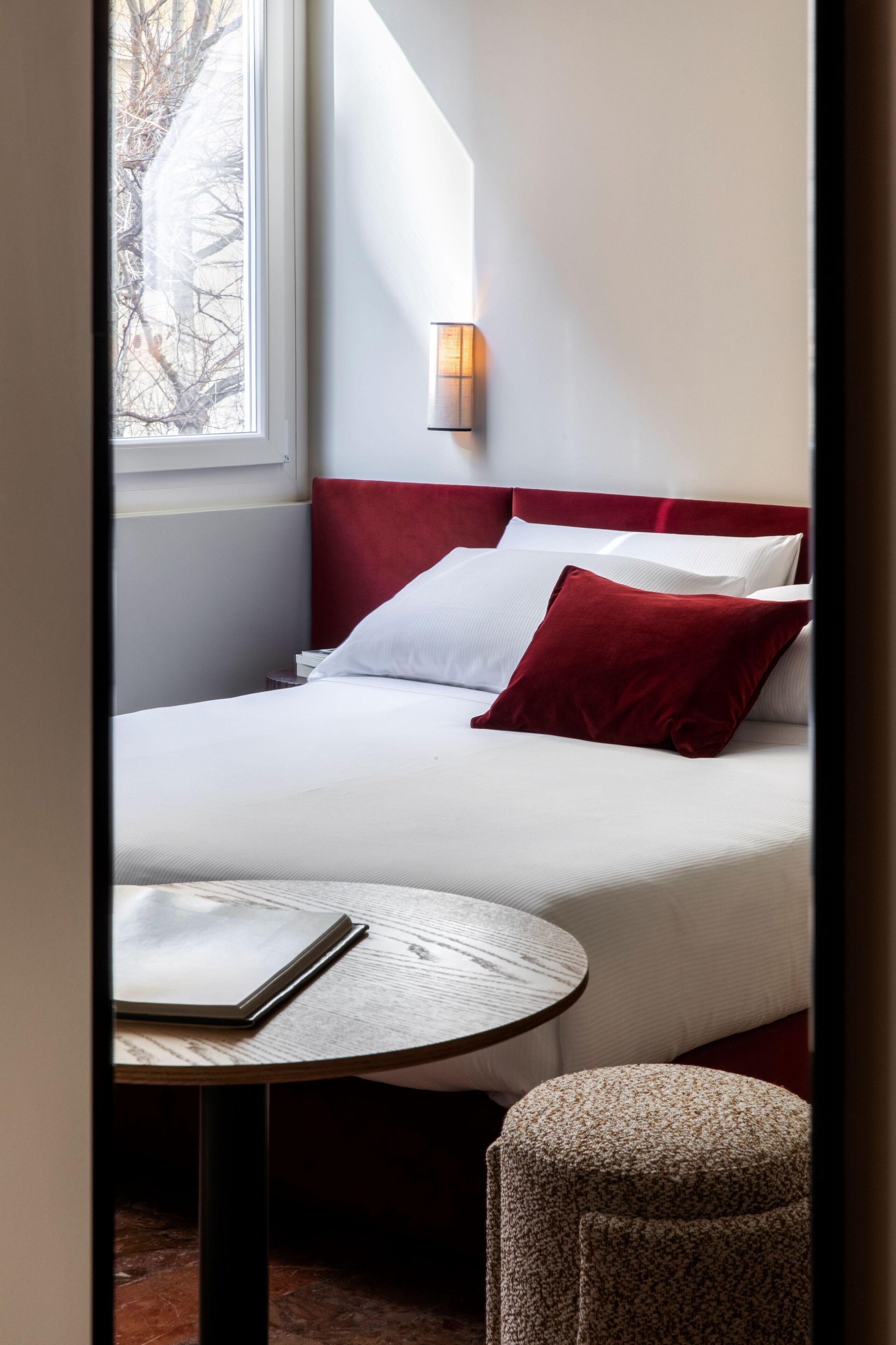 A hotel room with a bed, a bedside table, a chair, and a window with a view of trees.