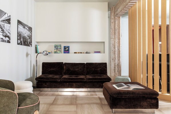 A modern living room with a dark brown sofa, a white armchair, and a brown ottoman with an open book. There are framed pictures on the wall and a floor lamp. - Atel Guest House (Rome)
