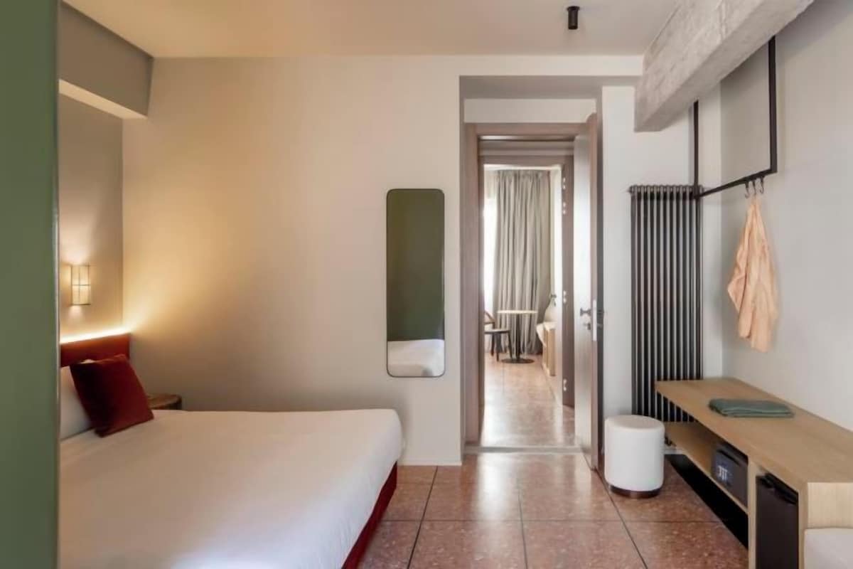 A hotel room with a bed, a bedside table, a mirror, a radiator, and a view into the dining area.
