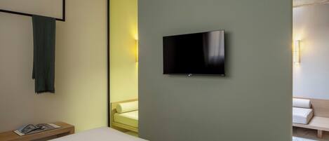 A modern hotel room with a bed, a bedside table with a book, a flat-screen TV mounted on the wall, and a seating area with a sofa and a bench.
