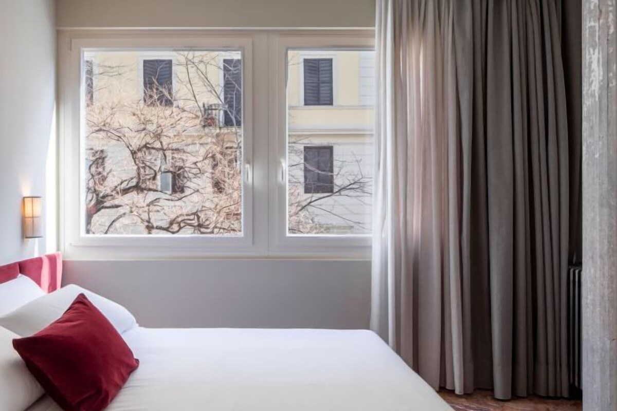 A hotel room with a bed, a window with a view of buildings, and a curtain.