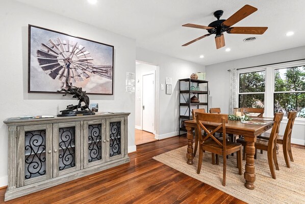 A dining area with a wooden table and chairs, a ceiling fan, and a large windmill painting on the wall. - Pine Street House - Close to Main (Fredericksburg)