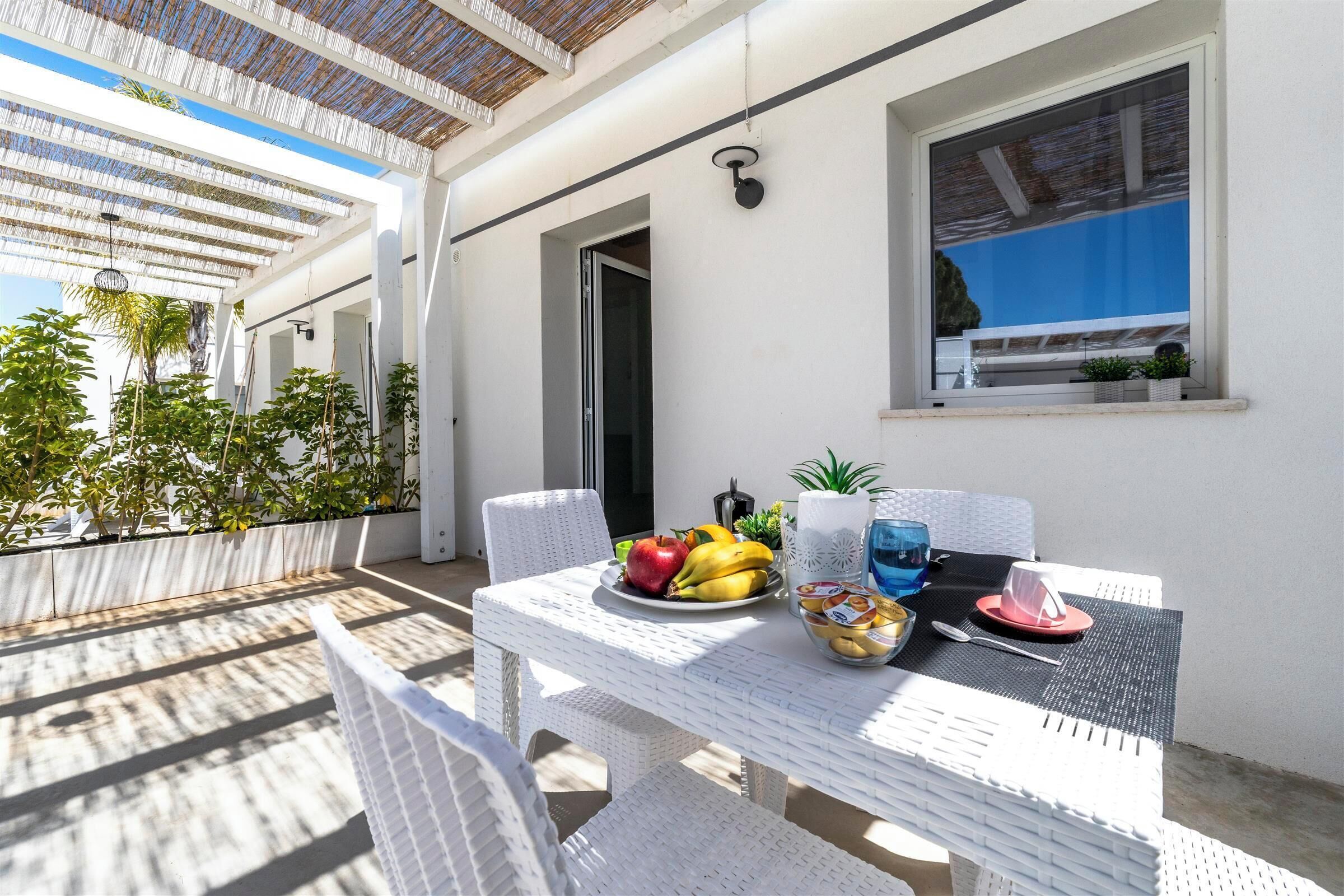 A white wicker table with a fruit bowl, a glass of water, and a red hat on a balcony with a white wall and a window.