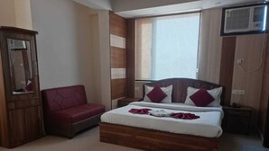 A bedroom with a bed, a red sofa, a wooden wardrobe, and a window with blinds. - Hotel Vistara Grand (Haridwar)