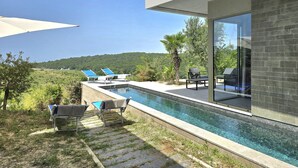 Un área de piscina moderna al aire libre con sillones de descanso y vistas a una colina boscosa.