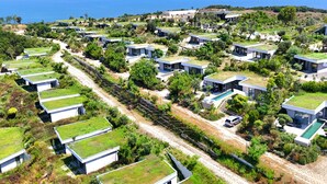 Un complejo turístico costero con edificios modernos y ecológicos de techos planos, dispuestos en línea a lo largo de un camino de tierra con vista al mar.