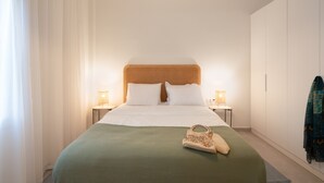 Et hotelværelse med en seng, to sengeborde, en lampe, et grønt dynedække, en hvid pude, et træhovedgærde, et ophængt tøjstykke og en lille håndtaske.