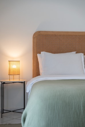 Un letto con lenzuola bianche, una testiera in vimini e un comodino con una lampada.