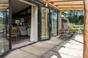 A modern outdoor patio with a wooden deck, a glass door leading to a kitchen area, and a dining table set for two.