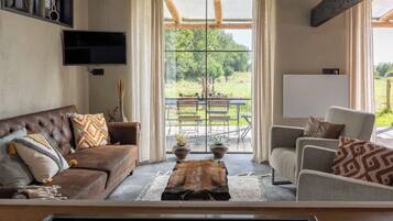 A living room with a brown leather sofa, a grey armchair, and a wooden coffee table. A television is mounted on the wall, and there is a large window with a view of a garden and outdoor seating.