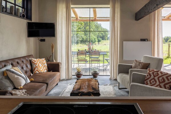 A living room with a brown leather sofa, a grey armchair, and a wooden coffee table. A television is mounted on the wall, and there is a large window with a view of a garden and outdoor seating.