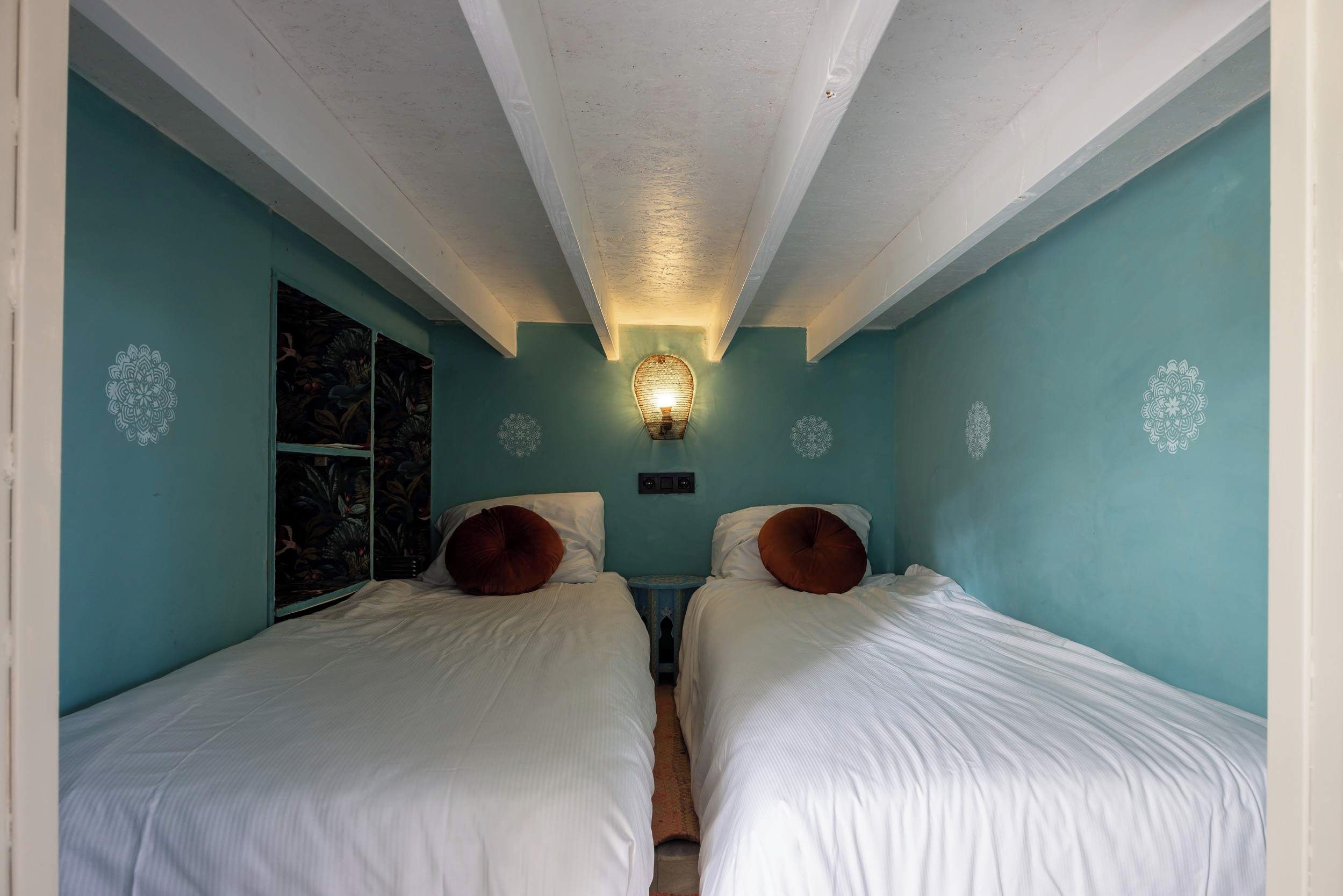 Ein Zimmer mit zwei Betten, einer Wandleuchte und einem Fenster mit Blumenmuster.