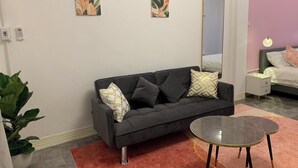 A modern living room with a grey sofa, a glass coffee table, and a pink rug. There are two wall-mounted paintings, a bed with a pink headboard, and a bedside table with a lamp. - Mana restaurant and boutique hotel (Khon Kaen)