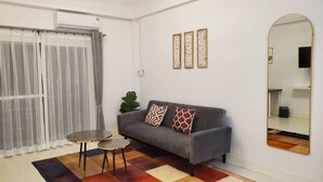 A living room with a grey sofa, two patterned cushions, a small round table, and a colorful rug. There is a large mirror on the wall and a potted plant beside the sofa. - Mana restaurant and boutique hotel (Khon Kaen)