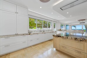A modern kitchen with white cabinetry, marble countertops, and large windows offering a view of the outdoors.