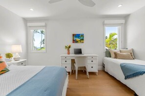 A bedroom with a bed, desk, and chair, a window with a view of palm trees, and a painting on the wall.