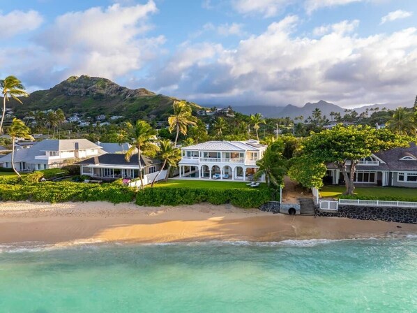 A beachfront property with a large house, a sandy beach, and a mountain in the background.