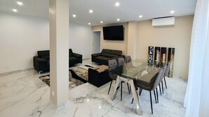A modern living room with a glass dining table, black sofas, and a flat-screen TV mounted on the wall. - L'Exclusif - Paris - Neuilly - Levallois (Levallois-Perret)