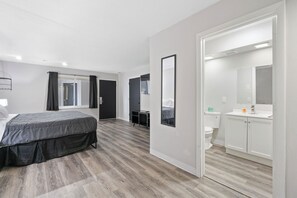 A modern hotel room with a bed, a small desk, a mirror, and a bathroom with a toilet and sink. - Willow Inn Suites (Eau Claire)