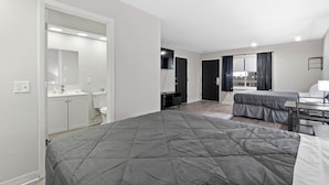 A modern hotel room with a large bed, a desk, a chair, a small table, a mirror, a window with curtains, and a bathroom with a sink and toilet. - Willow Inn Suites (Eau Claire)