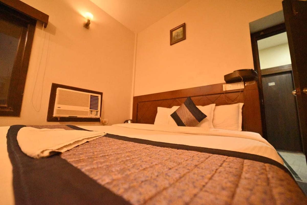 A hotel room with a bed, a window with blinds, an air conditioning unit, and a door.