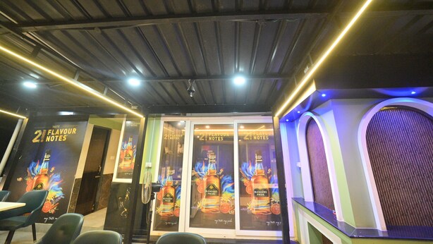A restaurant interior with a modern design, featuring a dark ceiling with exposed beams, illuminated by linear lights. There are promotional posters on the walls and a counter with a speaker. - Hotel Orion  (Bhadrak)