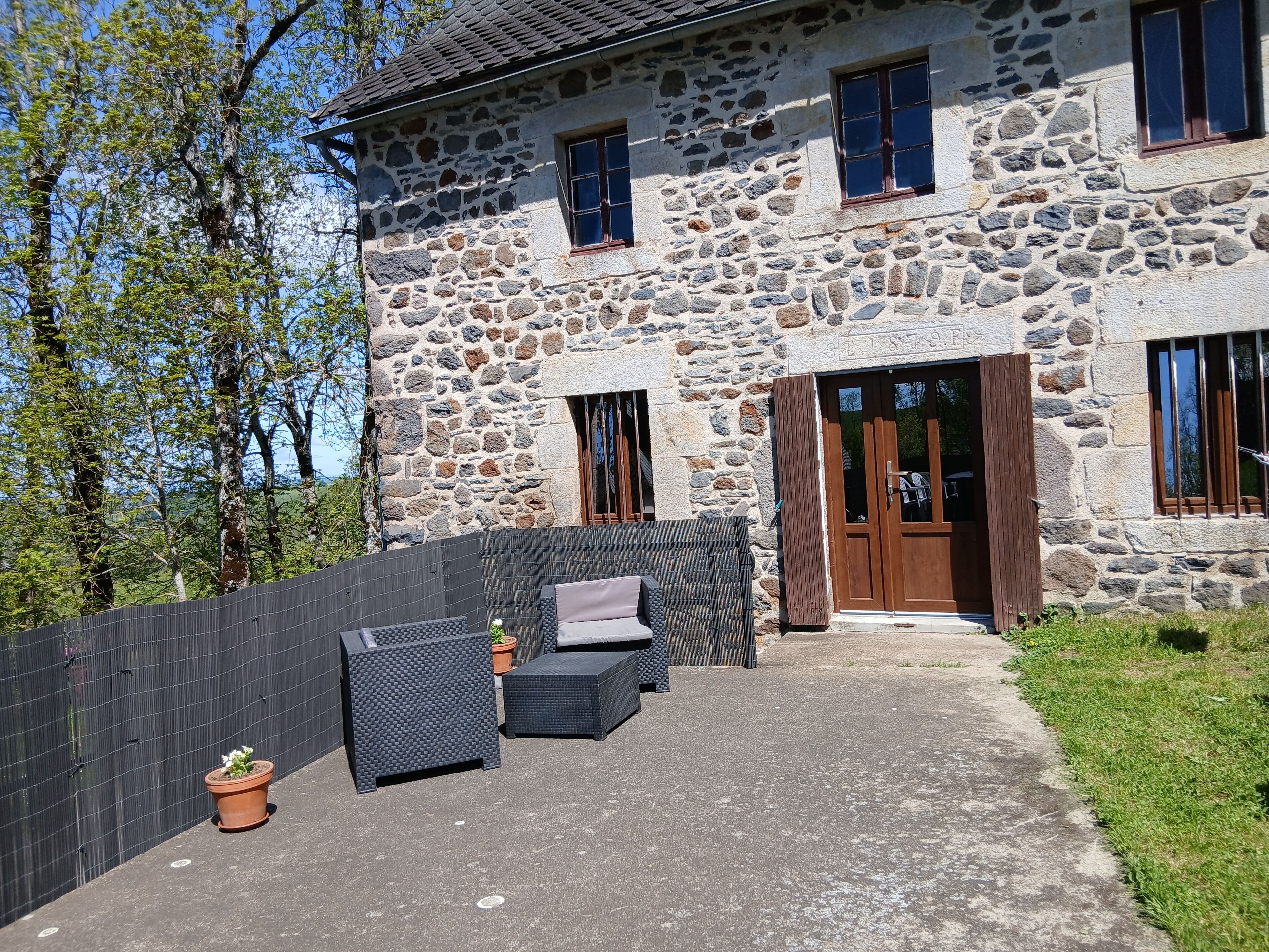 Un bâtiment en pierre avec une entrée pavée, une porte en bois et un petit jardin agrémenté de meubles d’extérieur et d’une plante en pot.
