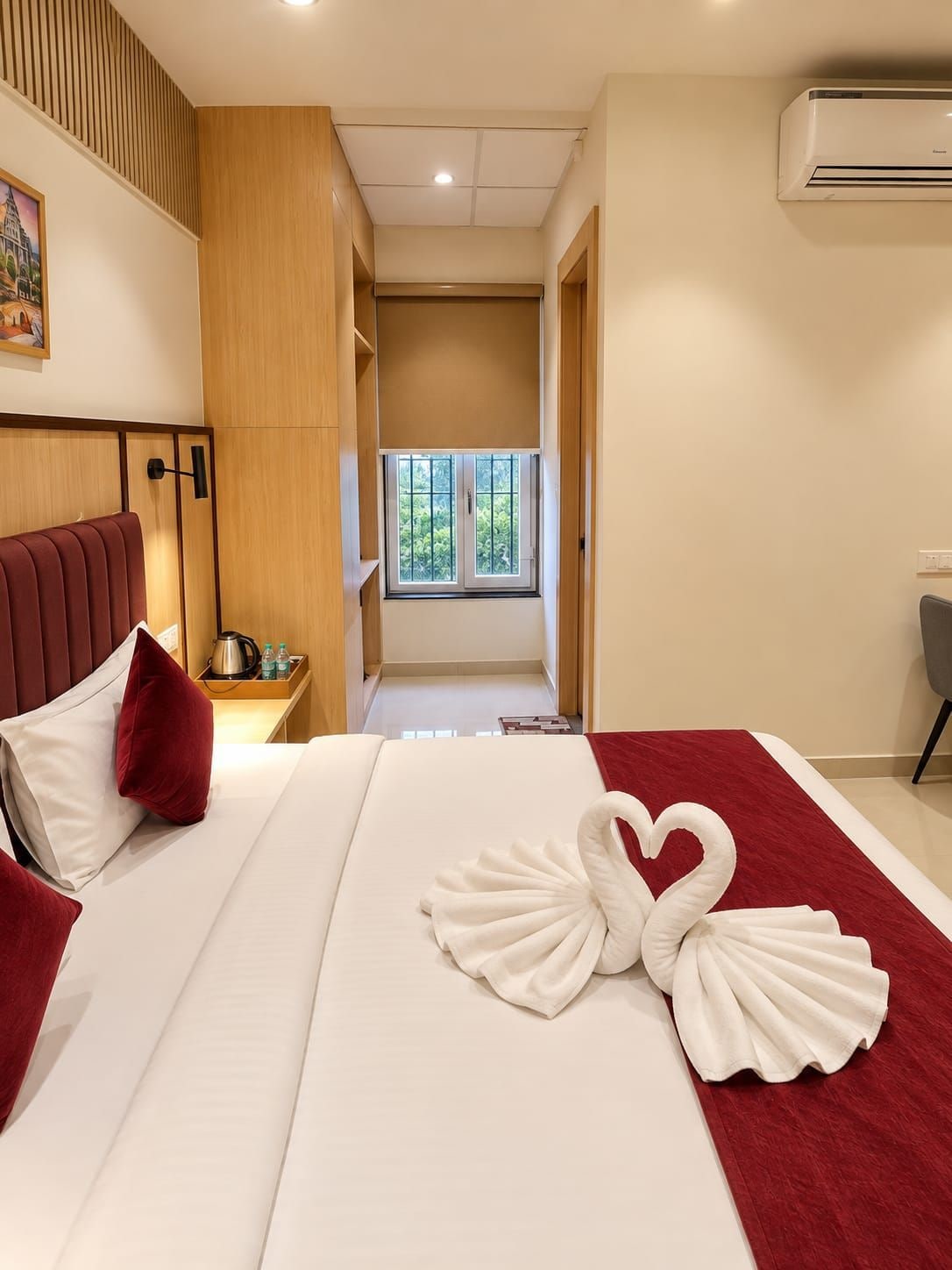 A hotel room with a bed, a red and white bedspread, a heart-shaped towel decoration, a window with a view, and an air conditioning unit.