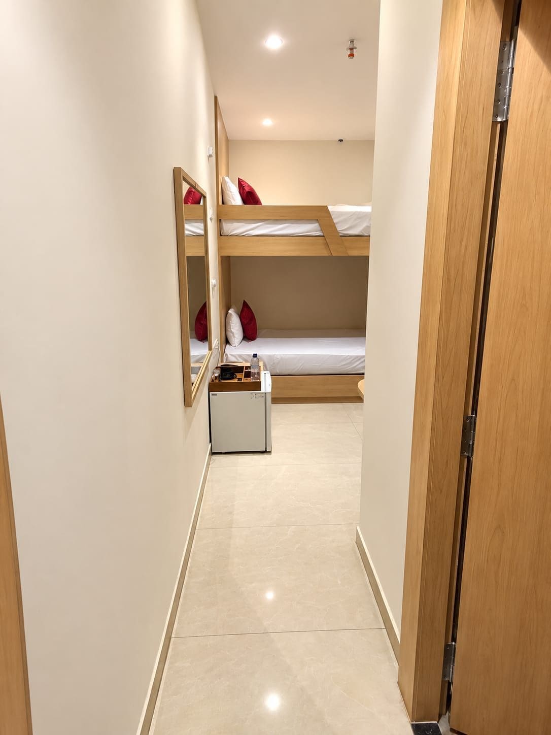 A narrow hallway leading to a bunk bed room with a small refrigerator and a mirror.