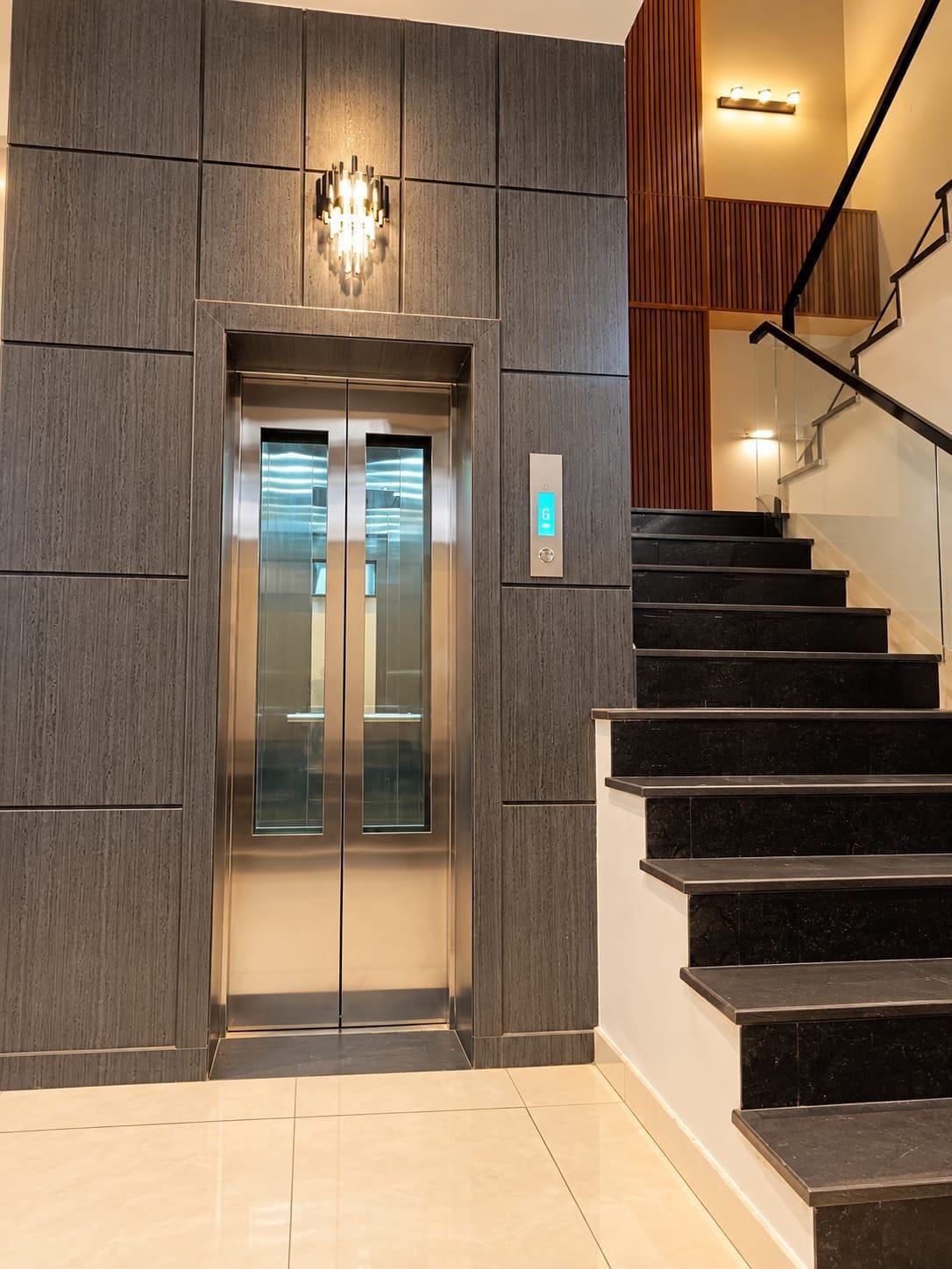 A modern elevator with a digital display, a staircase with black steps, and a wall-mounted light fixture.