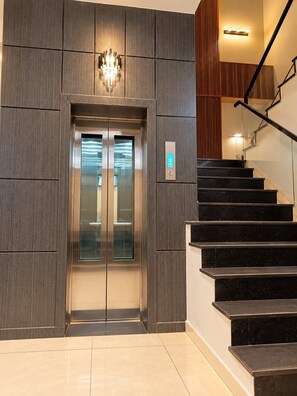 Un ascenseur moderne doté d’un écran numérique, un escalier avec des marches noires et un luminaire fixé au mur.