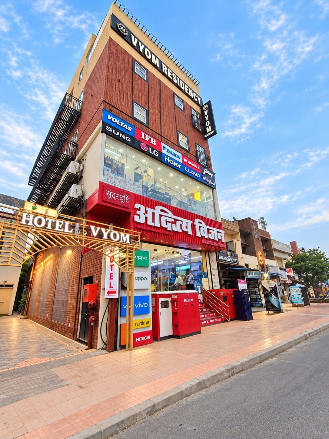 A hotel with a prominent 'HOTEL VYOM' sign, multiple brand stores on the ground floor, and a clear sky above.