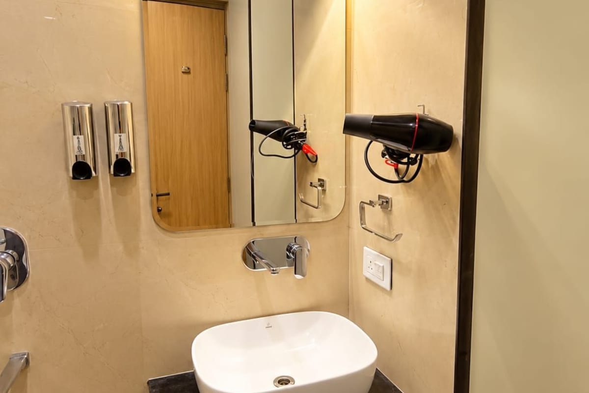 Un baño con lavamanos blanco, espejo, secador de pelo y cubo de basura.