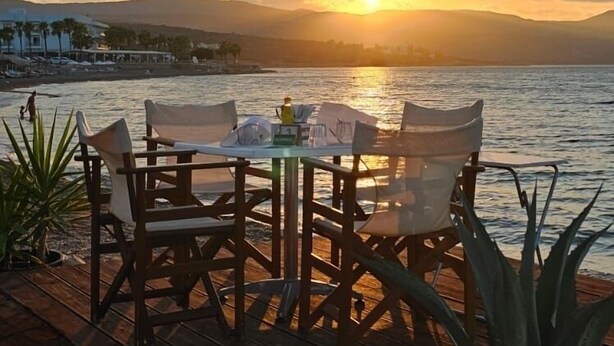 Eine Holzterrasse mit Tisch und Stühlen, die einen Sonnenuntergang über dem Meer zeigt.