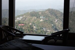 Una habitación con cama, dos sillas y un ventanal que ofrece vistas a un paisaje montañoso con edificios.