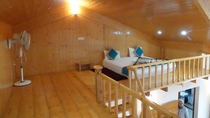 Eine Holzhütte mit Schlafzimmer im Zwischengeschoss, einem Bett mit blauen Kissen, einem Standventilator und einer Treppe.
