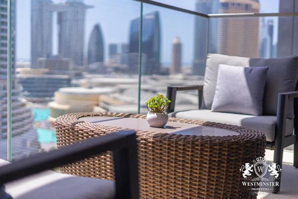 A modern lounge with a wicker chair, a small table with a plant, and a sofa with cushions. A city skyline is visible through large windows.