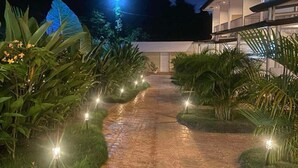 A paved pathway leading to a modern building with a glass facade, surrounded by lush greenery and illuminated by ground lights. - DMC Condotel (Puerto Princesa)